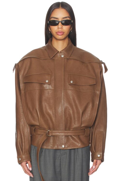 Model wearing a fully zipped and buttoned brown leather bomber jacket featuring an adjustable buckled waist belt, sharp collar, and structured flap pockets.