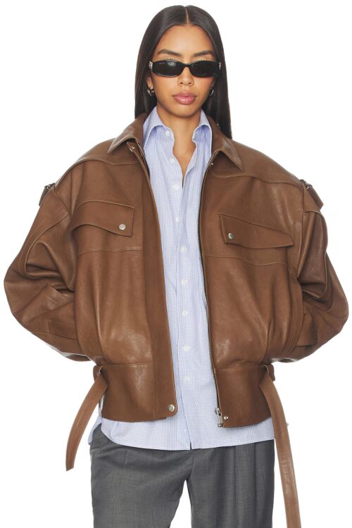 Model wearing an open brown leather bomber jacket with broad shoulder epaulettes, chest flap pockets, and unfastened waist belt straps styled over a light blue collared shirt.