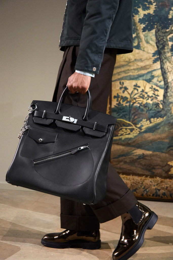 Hermès Black Leather Tote Handbag with Silver-Tone Hardware – Luxury Lifestyle Image Person carrying a premium black leather tote handbag with silver-tone hardware, front utility pocket, zipper compartment, dual top handles, and a detachable chain accessory in an elegant indoor setting.