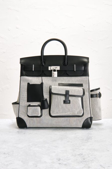 Structured cream canvas and black leather multi-pocket travel tote standing upright on a light stone floor against a textured plaster wall.