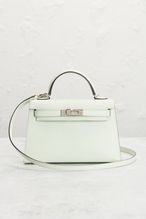 A light mint green textured calfskin mini handbag displayed upright on a light stone surface against a textured plaster wall, showcasing its silver turnlock plate and wrapped detachable leather strap.