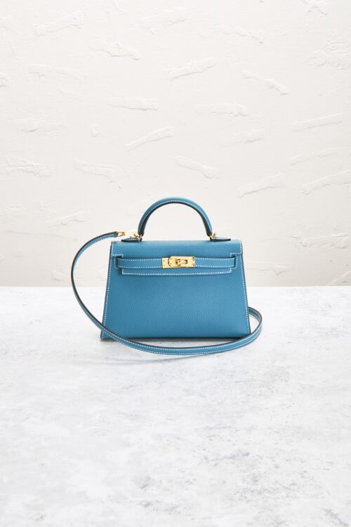 Front perspective of a mini blue leather handbag sitting on a textured grey counter with a subtle white wall backdrop, showing its top handle and gold turnlock clasp.