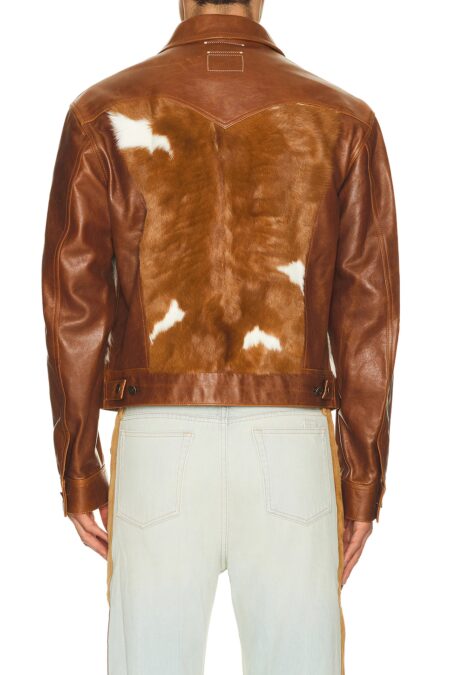 Back view of a model wearing a brown leather trucker jacket, displaying a large brown and white spotted cow fur back panel with Western yoke stitching details.