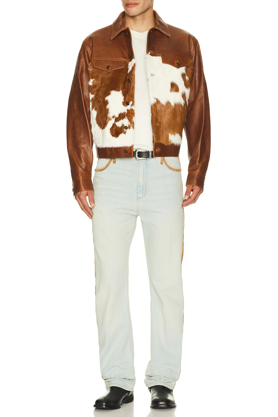 Full length portrait of a model wearing a brown leather trucker jacket featuring cow fur front panels, paired with light blue jeans and black boots against a studio background.