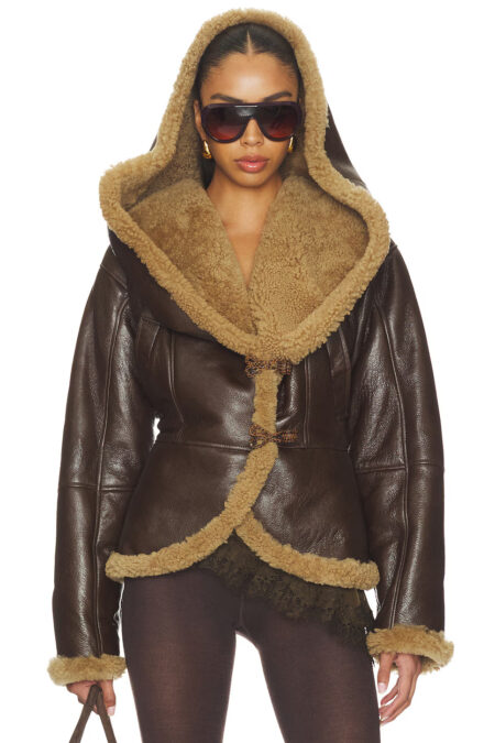 A model wearing a dark brown leather shearling hooded jacket with tan lining, oversized hood, and loop closures, styled with sunglasses and gold earrings