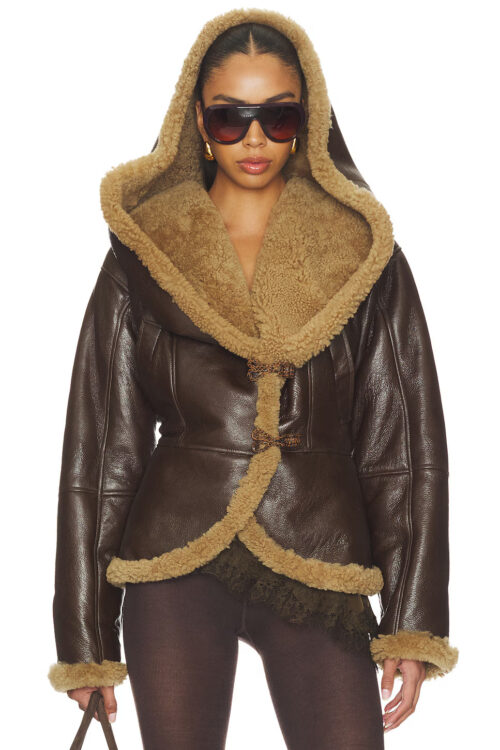 A model wearing a dark brown leather shearling hooded jacket with tan lining, oversized hood, and loop closures, styled with sunglasses and gold earrings
