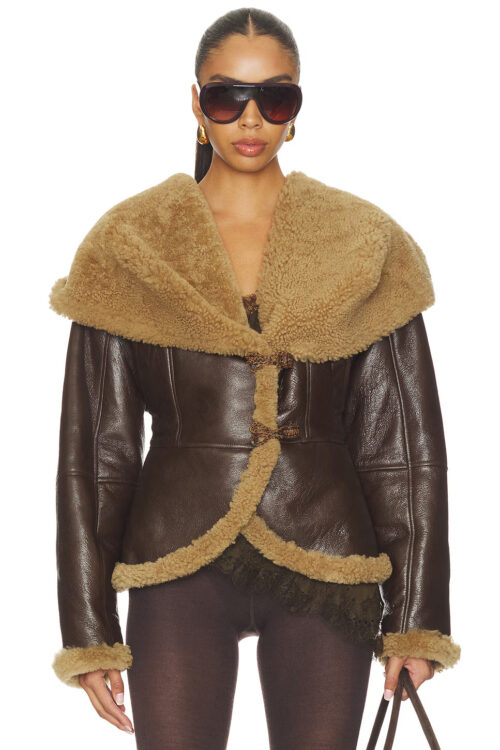 A model wearing a dark brown leather shearling hooded jacket with tan lining, oversized hood, and loop closures, styled with sunglasses and gold earrings.
