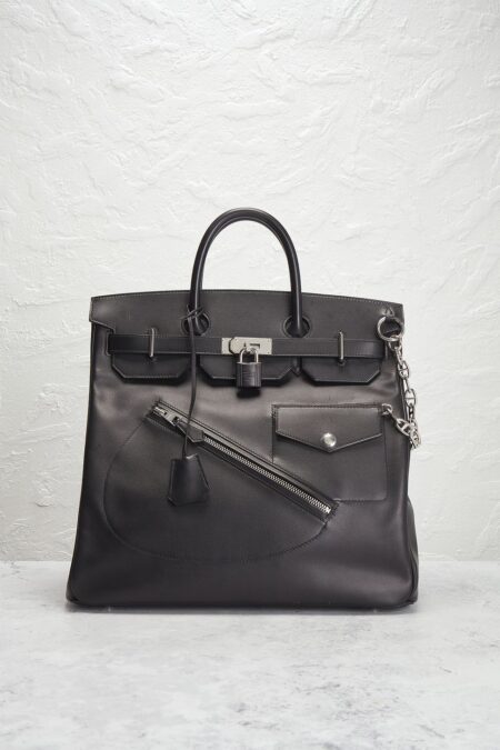 A structured black calfskin leather travel bag with a metal lock and diagonal front zipper details, resting upright against a textured off-white wall background.