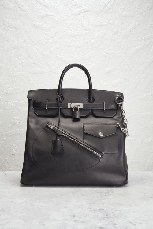 A structured black calfskin leather travel bag with a metal lock and diagonal front zipper details, resting upright against a textured off-white wall background.