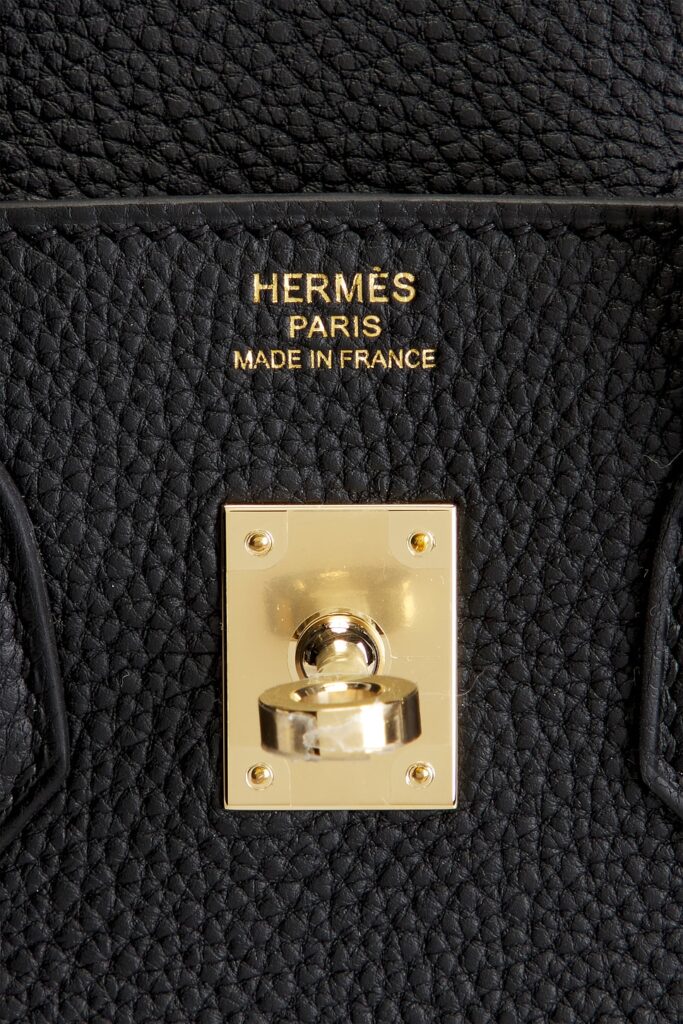 Macro close-up of a metallic gold foil heat stamp reading words of origin over a gold turn-lock plate on premium black pebbled leather.