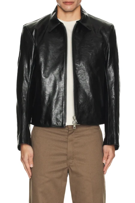 A front-facing view of a model wearing a structured black heavyweight leather jacket featuring a classic pointed collar and a dual front zipper closure slightly unzipped at the hem, paired with light brown trousers.