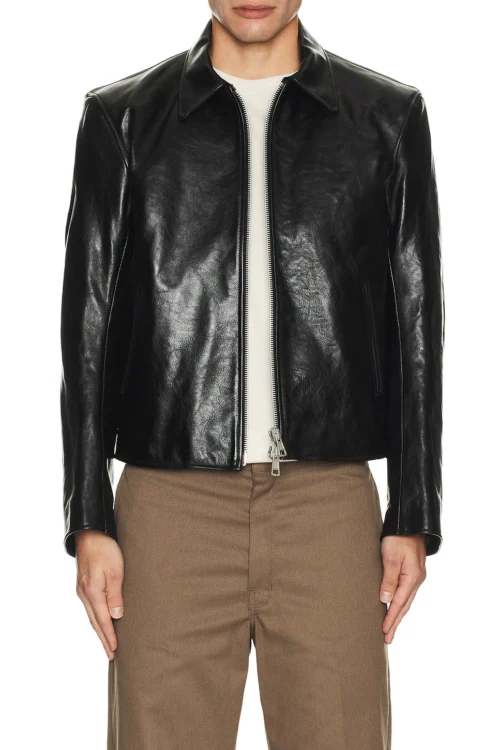 A front-facing view of a model wearing a structured black heavyweight leather jacket featuring a classic pointed collar and a dual front zipper closure slightly unzipped at the hem, paired with light brown trousers.
