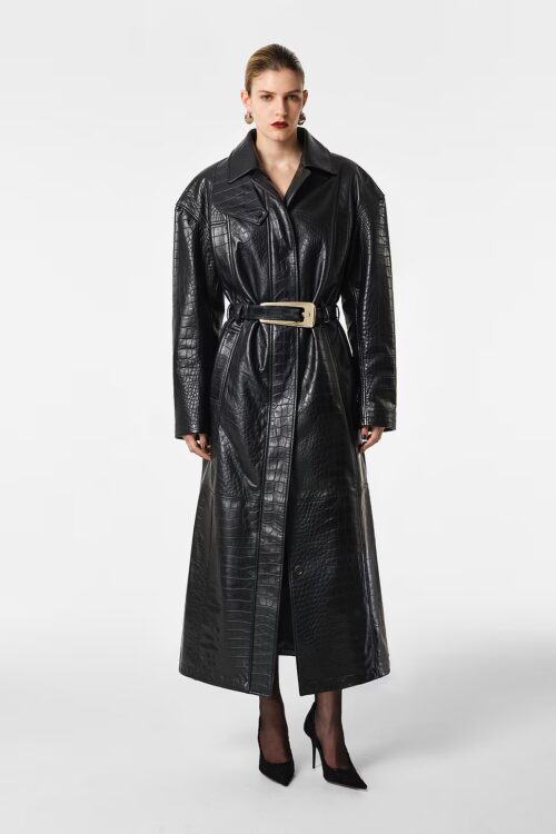 Full-length front view of a model wearing an ankle-length black croc-embossed leather coat cinched at the waist with a gold buckled belt, styled with black tights and pointed high heels.