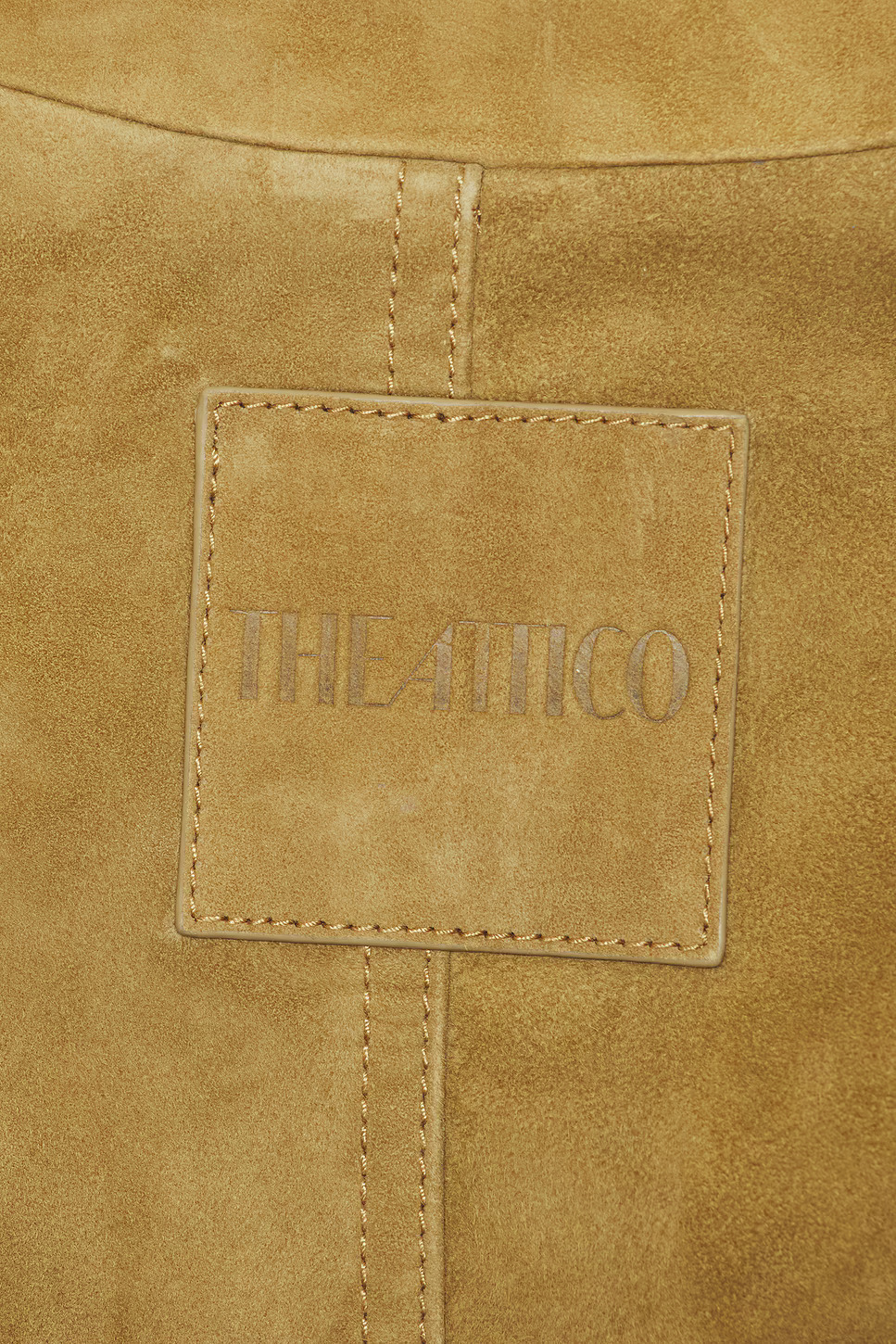 Macro detail shot focusing on the stitched square patch with debossed designer text on a textured tan goatskin suede surface.