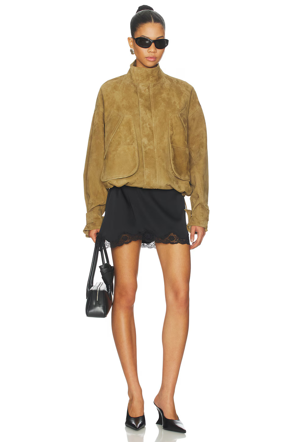 Full-length style shot of a model wearing an oversized brown suede bomber coat with a black miniskirt, matching black pumps, and a luxury handbag.