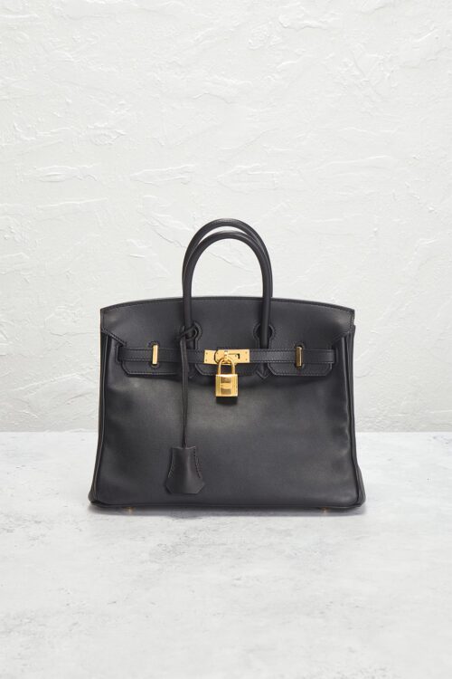 Front view of a compact black leather top-handle handbag resting on a grey textured counter with gold hardware accents and hanging key clochette.