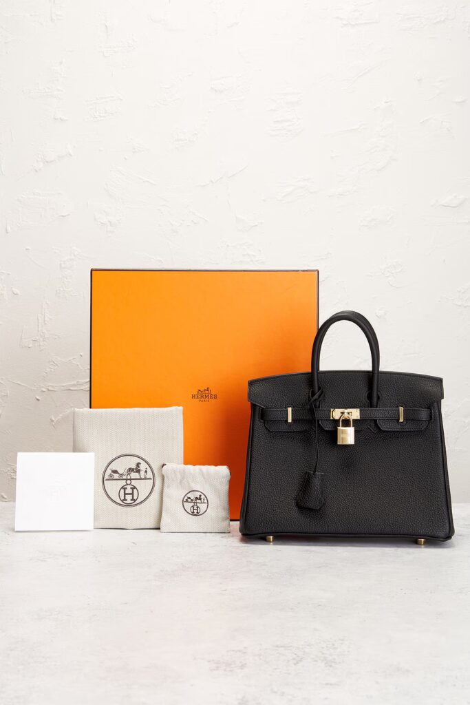 A black grained leather handbag displayed alongside its signature bright orange packaging box, two canvas dustbags, and a white booklet.