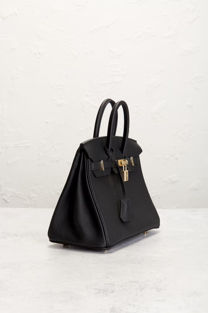 A three-quarter side angle of a black grained leather top-handle bag, showcasing its classic flared accordion silhouette and depth on a light surface.