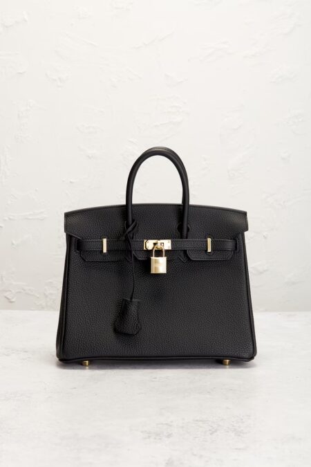 A mini black grained leather tote bag resting upright on a smooth stone floor against a rustic textured white wall, showcasing gold hardware and a matching leather clochette.