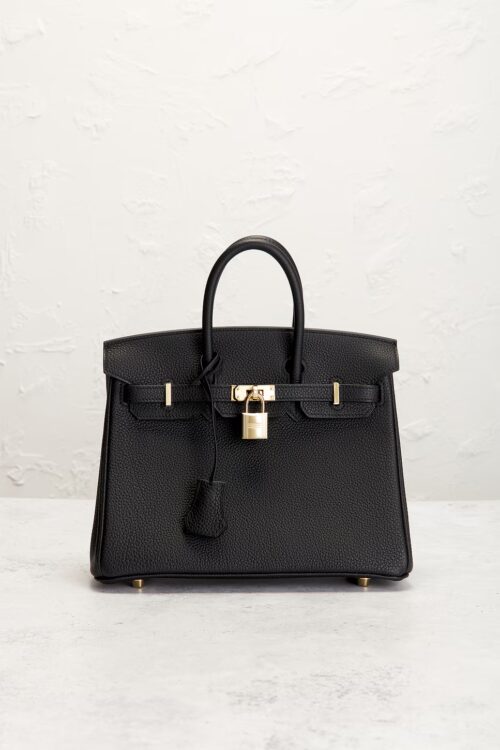 A mini black grained leather tote bag resting upright on a smooth stone floor against a rustic textured white wall, showcasing gold hardware and a matching leather clochette.