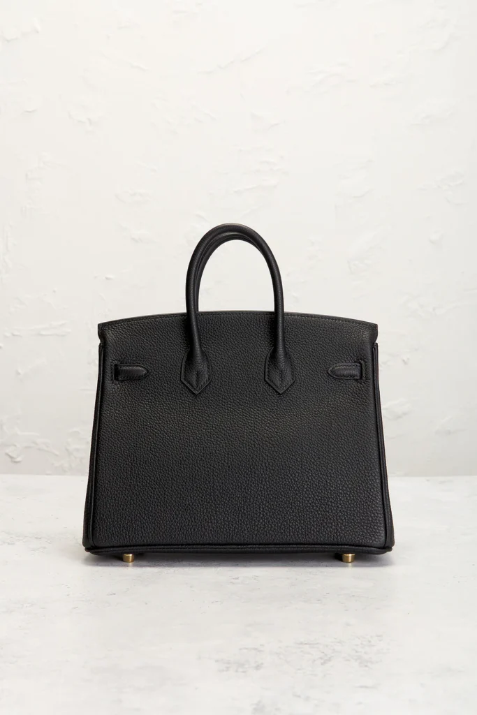 The rear view of a structured black pebbled leather handbag, highlighting the clean leather panels, structural stitching, and the base of the dual top handles.