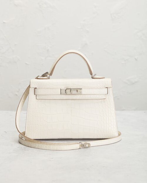 Front-facing white matte alligator leather handbag displayed in a lifestyle setting with structured top handle, detachable shoulder strap, polished silver-tone hardware, and elegant luxury styling.