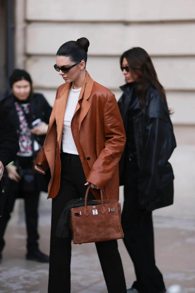 Women Luxury Designer Bags Collection | Premium Handbags at REVOLVE COLLECTION Elegant woman carrying a premium brown luxury designer handbag while wearing a sophisticated leather blazer, representing timeless women's luxury fashion accessories.