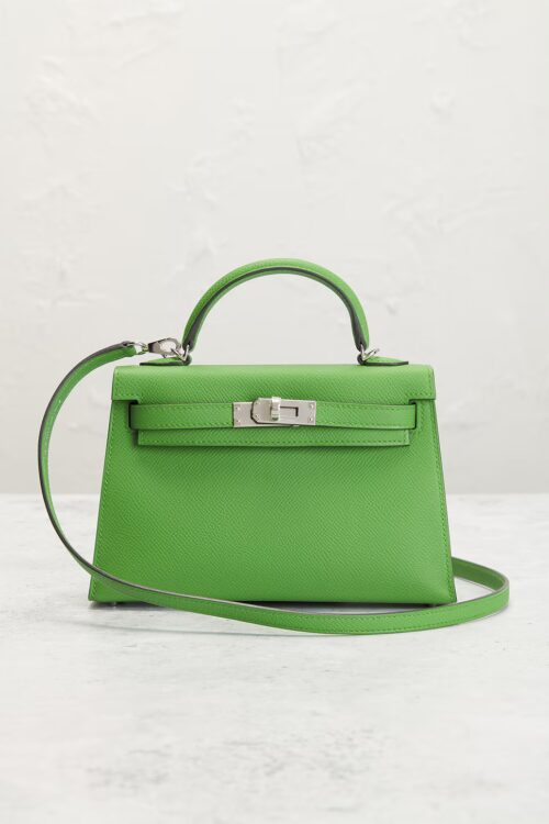 Front studio portrait of a bright green textured leather top-handle mini bag resting on a light grey stone surface, complete with silver sangles turnlock clasp and attached shoulder strap.