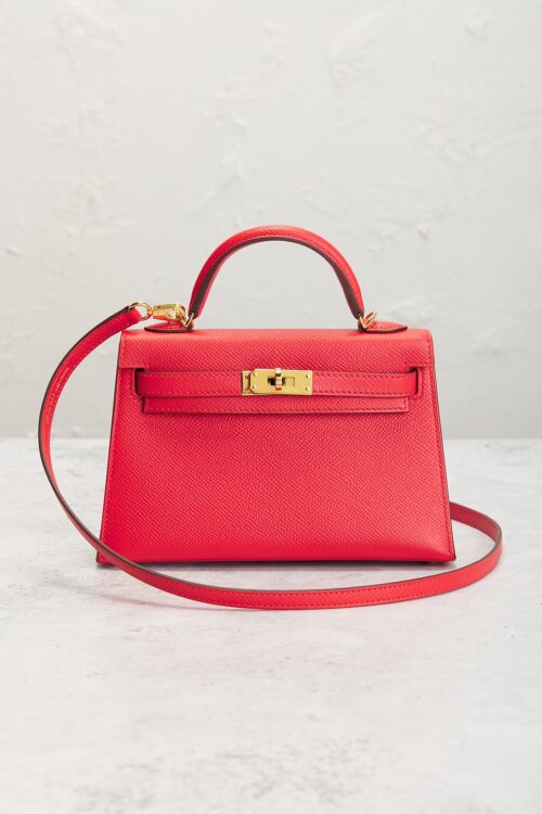 Front perspective of a bright pink mini leather top-handle bag resting on a textured light surface, showcasing its sharp Sellier edge stitching, gold sangles turnlock clasp, and curved shoulder strap.
