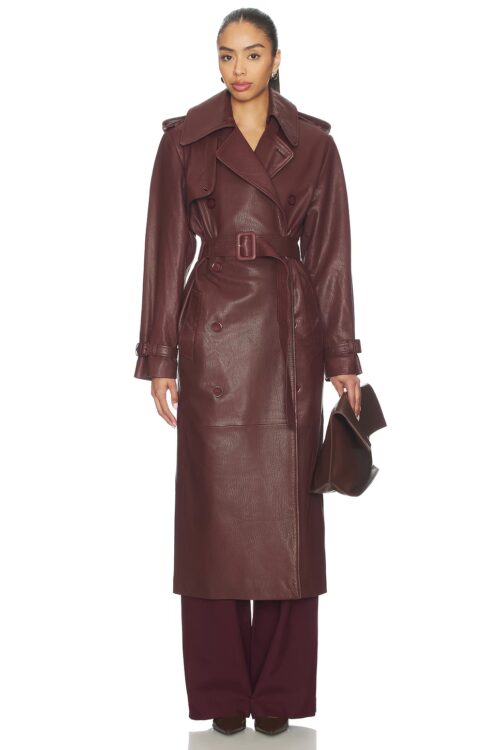 Full-length studio front view of a model wearing an ankle-length burgundy leather trench coat styled fully buttoned with a matching waist buckle belt, notched lapels, cuff tabs, and holding a dark leather clutch.