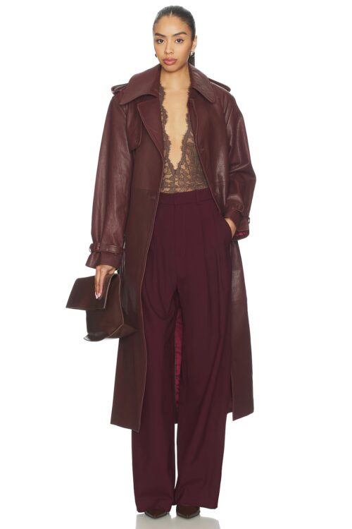 Full-length studio portrait of a model wearing an open burgundy leather trench coat draped over a sheer lace bodysuit and matching high-waisted wide-leg trousers.