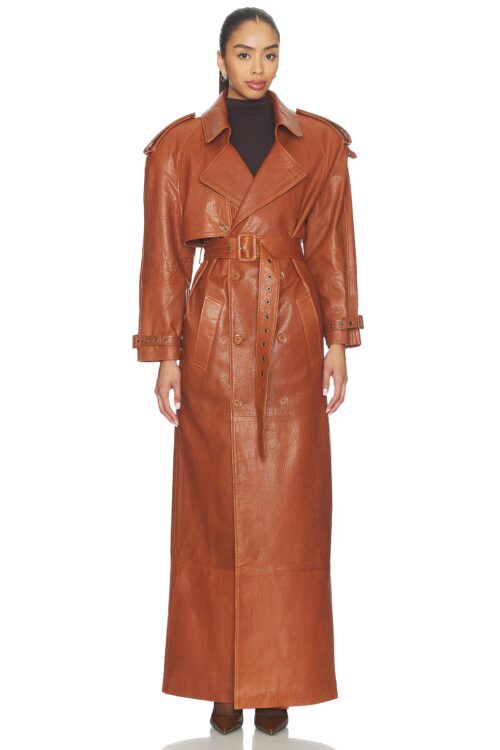 Full-length studio front view of a model wearing an ankle-length tan cognac leather trench coat with padded shoulders, notched lapels featuring silver-tone buckle detailing, and a cinched grommet buckle belt.