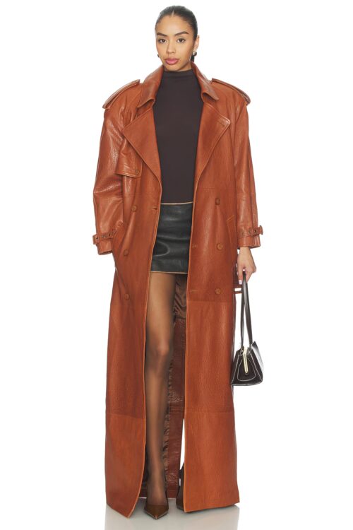 Full-length studio portrait of a model wearing an open cognac brown leather trench coat styled over a dark top, black leather mini skirt, brown pumps, and carrying a structured dark leather handbag.